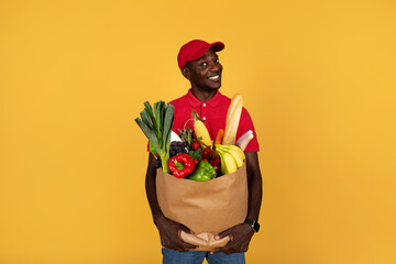 Curious smiling millennial black guy courier hold paper bag with products, look at free space