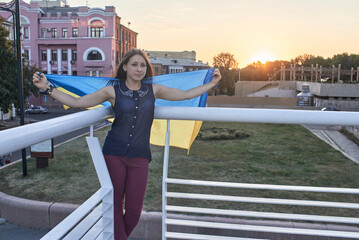 Photo of a Ukrainian woman holding a Ukrainian flag in her hands behind her back, she stands against the backdrop of the city. Sunset.