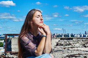 A girl who is not afraid of heights, with a calm face, sits next to a precipice against the backdrop of the city and the blue cloudy sky.