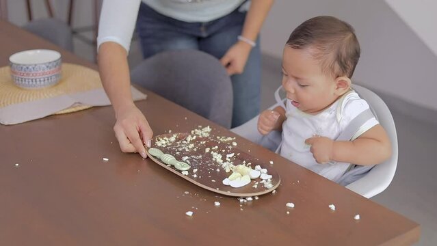 Baby trying his first solid foods, sitting at the table eating while his mother helps him. Baby led weaning concept