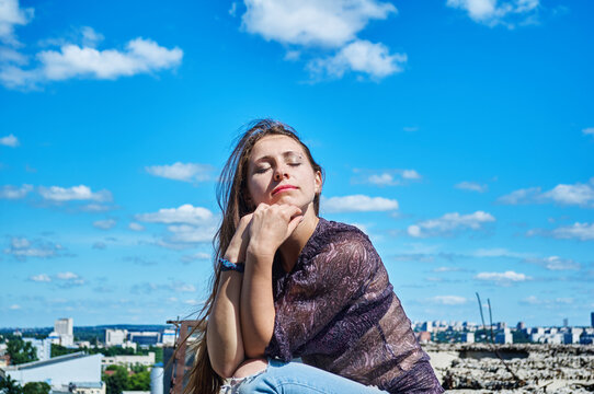 A girl who is not afraid of heights, with a calm face, sits next to a precipice against the backdrop of the city and the blue cloudy sky.