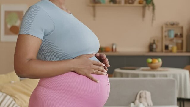Tilt up slowmo of young pregnant Black woman wearing tight pink leggings and crop t-shirt stroking her belly while practicing yoga at home in living room during leisure time