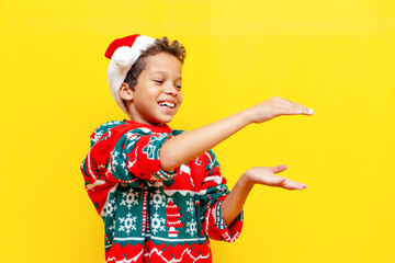 african american boy in christmas sweater and santa hat holds empty space with his hands on yellow...