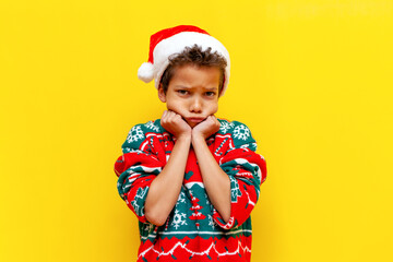 unhappy african american boy in a christmas sweater and santa hat is bored and sad on a yellow...