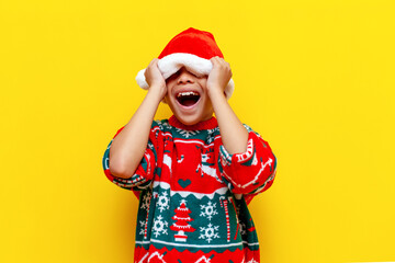 happy african american boy in christmas sweater and santa hat screams on yellow isolated background