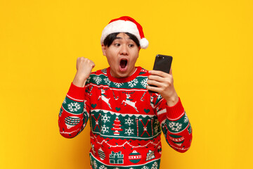 young asian man in christmas sweater and santa hat uses smartphone and celebrates victory