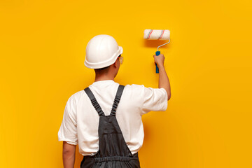 painter man in uniform works and paints the wall yellow with roller, repairman in hard hat and overalls