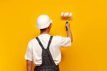 Foto op Plexiglas painter man in uniform works and paints the wall yellow with roller, repairman in hard hat and overalls © Богдан Маліцький