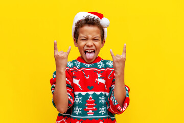 crazy african american boy in christmas sweater and santa hat shows tongue and rock gesture on yellow background