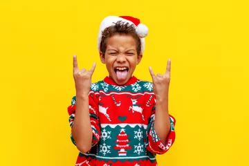 Foto op Canvas crazy african american boy in christmas sweater and santa hat shows tongue and rock gesture on yellow background © Богдан Маліцький
