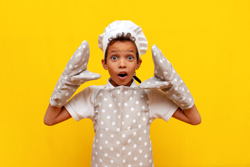 little boy african american baker in uniform and gloves looking surprised and looking at camera on yellow background - obrazy, fototapety, plakaty
