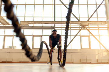 sporty african american man in sportswear trains in the gym in the morning, the guy waves the ropes...