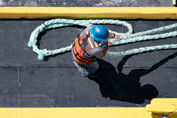 Overhead shot of a worker wearing a reflective vest and blue helmet, pulling a thick rope on a ship.