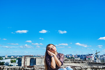 Fototapeta na wymiar A girl who is not afraid of heights, with a calm face, sits next to a precipice against the backdrop of the city and the blue cloudy sky.