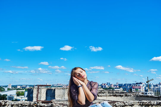 A girl who is not afraid of heights, with a calm face, sits next to a precipice against the backdrop of the city and the blue cloudy sky.