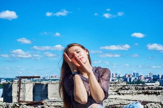 A girl who is not afraid of heights, with a calm face, sits next to a precipice against the backdrop of the city and the blue cloudy sky.