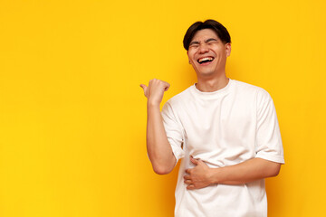 young asian guy in white t-shirt laughing and pointing to the side on yellow isolated background - obrazy, fototapety, plakaty