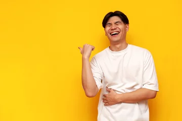 Fotobehang young asian guy in white t-shirt laughing and pointing to the side on yellow isolated background © Богдан Маліцький
