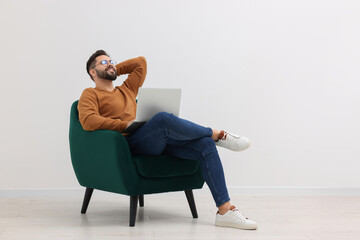 Handsome man with laptop relaxing in armchair near white wall indoors, space for text - obrazy, fototapety, plakaty