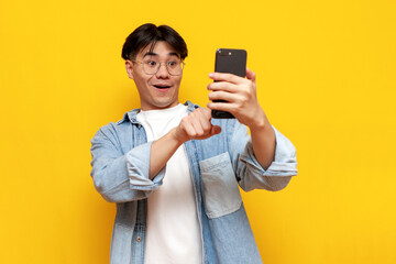 young asian guy with glasses uses smartphone on yellow isolated background, korean man selects...