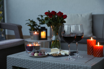 Glasses of wine, vase with roses, burning candles and snacks on rattan table at balcony in evening, closeup