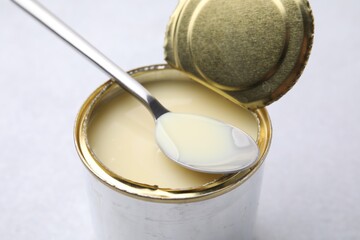 Open tin can with condensed milk and spoon on white table, closeup
