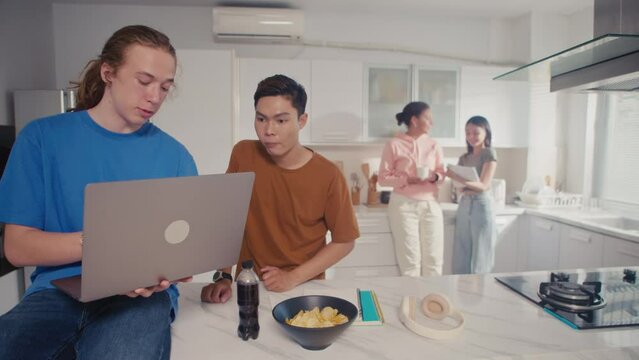 Medium long shot of young multi-ethnic men discussing homework using laptop while their male friends talking in background at kitchen