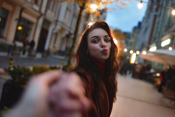 follow me. Beautiful romantic brunette girl in red coat takes her boyfriend's hands while walking in the evening city