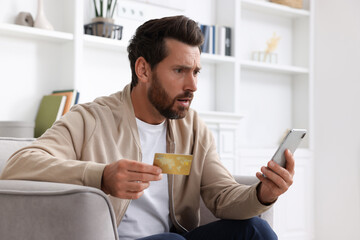 Upset man with credit card using smartphone in armchair at home. Be careful - fraud