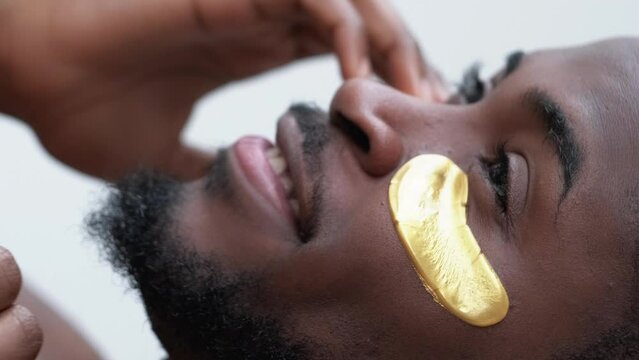 Vertical video. Male facial care. Skin rejuvenation. Handsome happy man with morning routine skincare gold collagen gel eye patch on perfect radiant face.