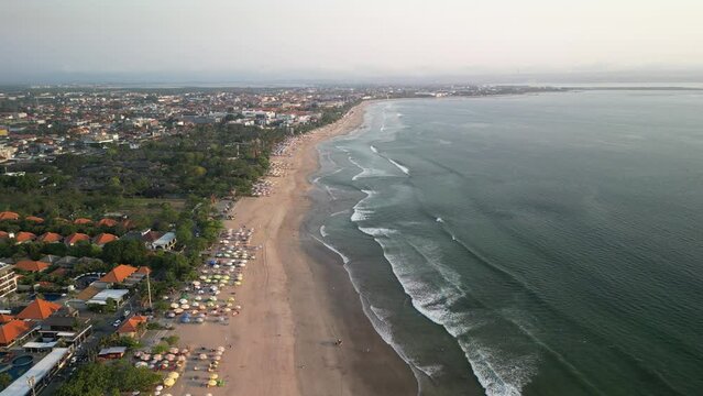 Aerial view of Beautiful sunset on the double six beach with Hotel, resort, cafe buildings, beach umbrella and calm ocean wave in Bali. Tourists are surfing, swimming and walking along the beach.