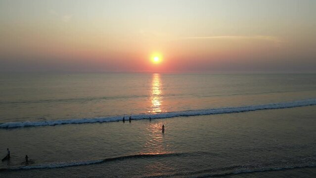 Aerial view of Tourists are enjoying the beautiful sunset on the beach with calm ocean wave in Bali. Tourists are surfing, swimming and walking on the beach at dusk with clear sky.