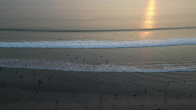 Aerial view of Tourists are enjoying the beautiful sunset on the beach with calm ocean wave in Bali. Tourists are surfing, swimming and walking on the beach at dusk with clear sky.