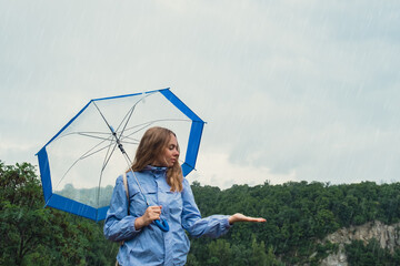 Blonde girl checking the weather under transparent umbrella outside in park. Autumn time rainy bad...