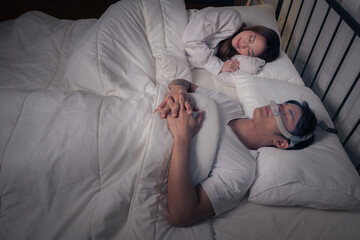Happy Asian couple with CPAP machine on side table with husband wearing CPAP mask sleeping smoothly in bed all night without snoring