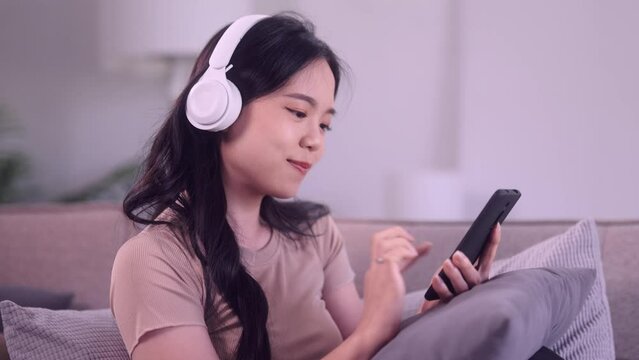 An Asian teenage girl uses her mobile phone to select her favorite songs, listens through headphones, and gently sways on a sofa in her living room, creating a cozy ambiance