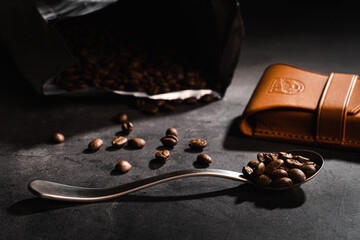 Coffee beans on a metal spoon and leather bag on dark concrete background