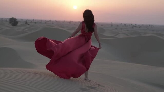 Young woman with black hair walking barefoot in a long red evening dress on the sand dunes of the desert
