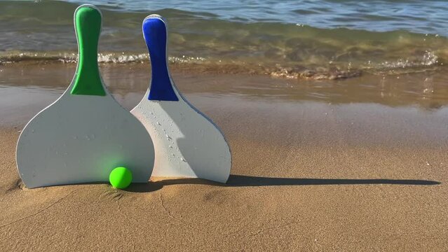 Pair of Beach tennis paddle rackets and green balls on sandy beach with waves breaking on shore. Sand for copy space