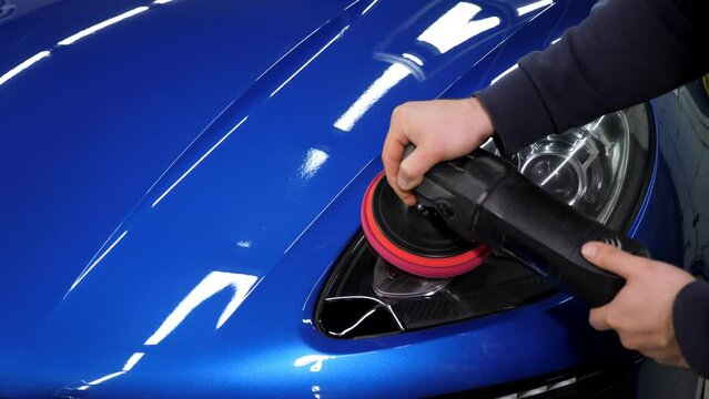 Close-up of a man polishing a headlight on a blue car with a polishing machine in a car dealership. The concept of detailing and polishing the car.