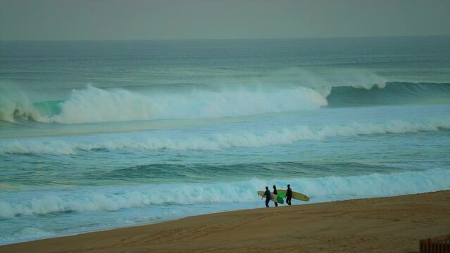 Huge swell big waves Hossegor Biarritz France view from beach shore two male surfers walking down sand looking at the surf getting ready to paddle in early morning cinematic slow motion follow pan
