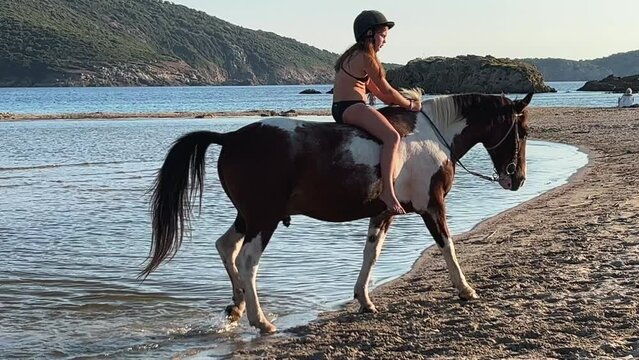 Young red-haired child girl in bikini have fun bathing and bareback riding horse in sea water in summer season