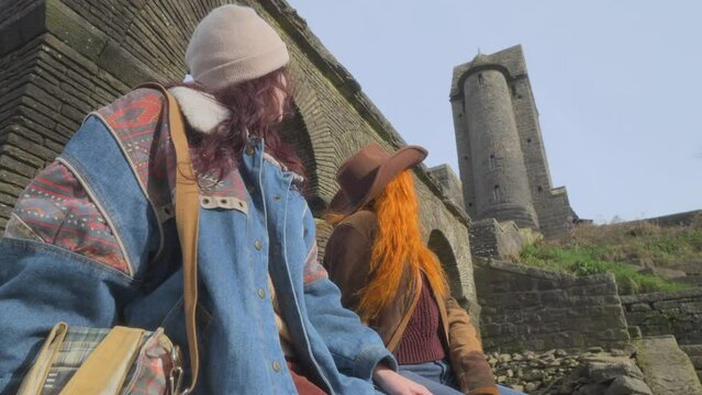 Young women, brunette and redhead resting and staring up at tall stone tower building. Half speed slow motion. Quarter stop pro mist filter.