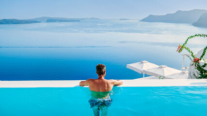 a young men in swim shorts relaxing in the pool looking out over the caldera of Santorini Island Greece, man at an infinity pool, a young guy on a luxury vacation in Europe Greece. - obrazy, fototapety, plakaty