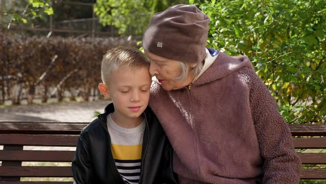 An old grandmother is sitting on a park bench with her little grandson, she hugs and kisses the boy. A great-grandmother with a child on a bench in autumn. Stable image.