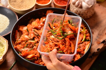  picks up kimchi in a plastic container with chopsticks from a bowl of shredded vegetables, including scallions, cabbage and carrots. There are rows of condiments, including Korean chilies. - obrazy, fototapety, plakaty