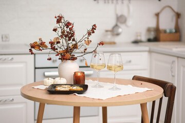 Autumn home interior in kitchen. Light dinner - backed pumpkin with cheese, glasse of white wine, burning candle, vase with dry branches on round wooden table in light kitchen.