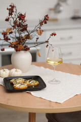 Autumn home interior in kitchen. Light dinner - backed pumpkin with cheese, glasse of white wine, burning candle, vase with dry branches on round wooden table in light kitchen.