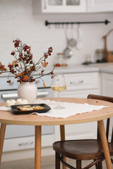 Autumn home interior in kitchen. Light dinner - baked pumpkin with cheese, glasse of white wine, burning candle, vase with dry branches on round wooden table in light kitchen.