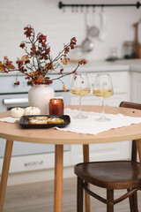 Autumn home interior in kitchen. Light dinner - baked pumpkin with cheese, glasse of white wine, burning candle, vase with dry branches on round wooden table in light kitchen.